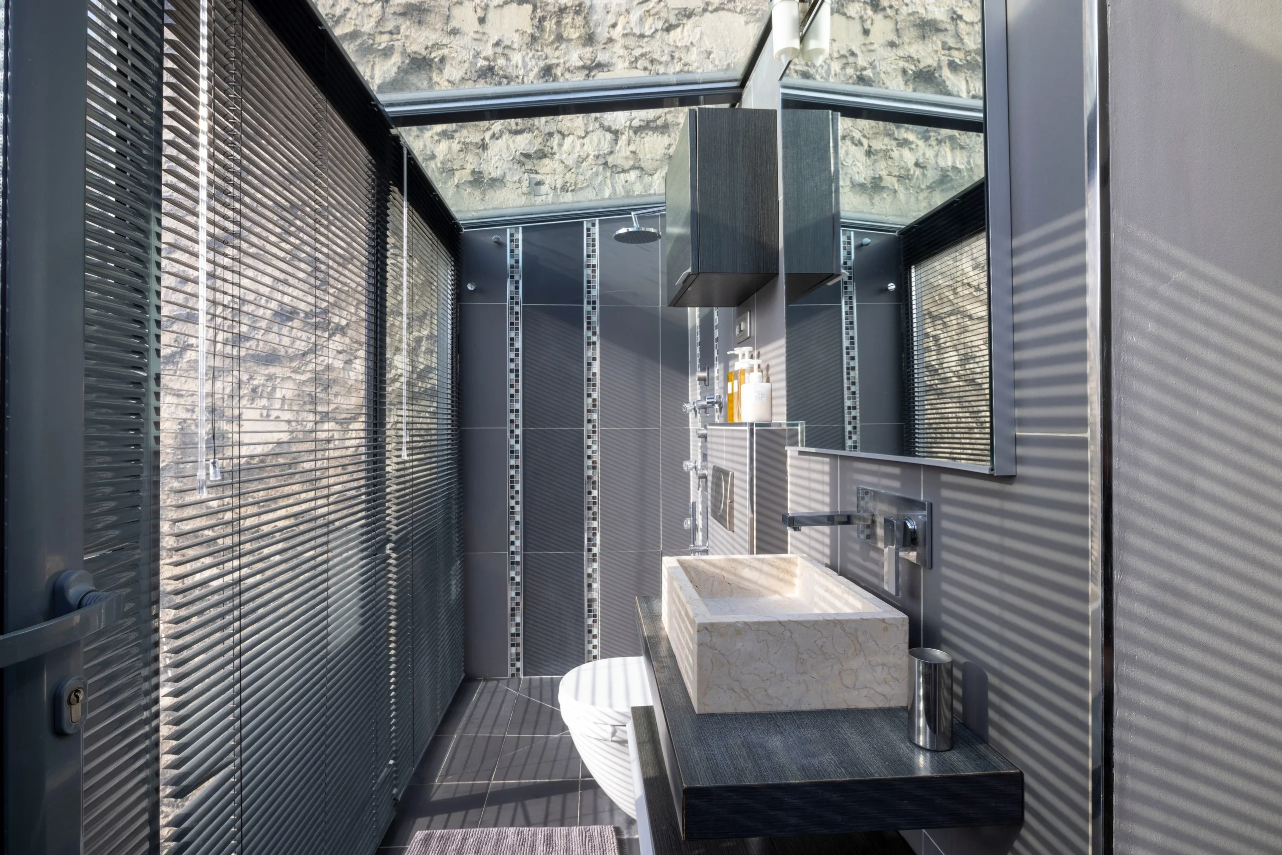 Salle de bains sous verrière avec douche, vasque rectangulaire en pierre claire et stores à lamelles.