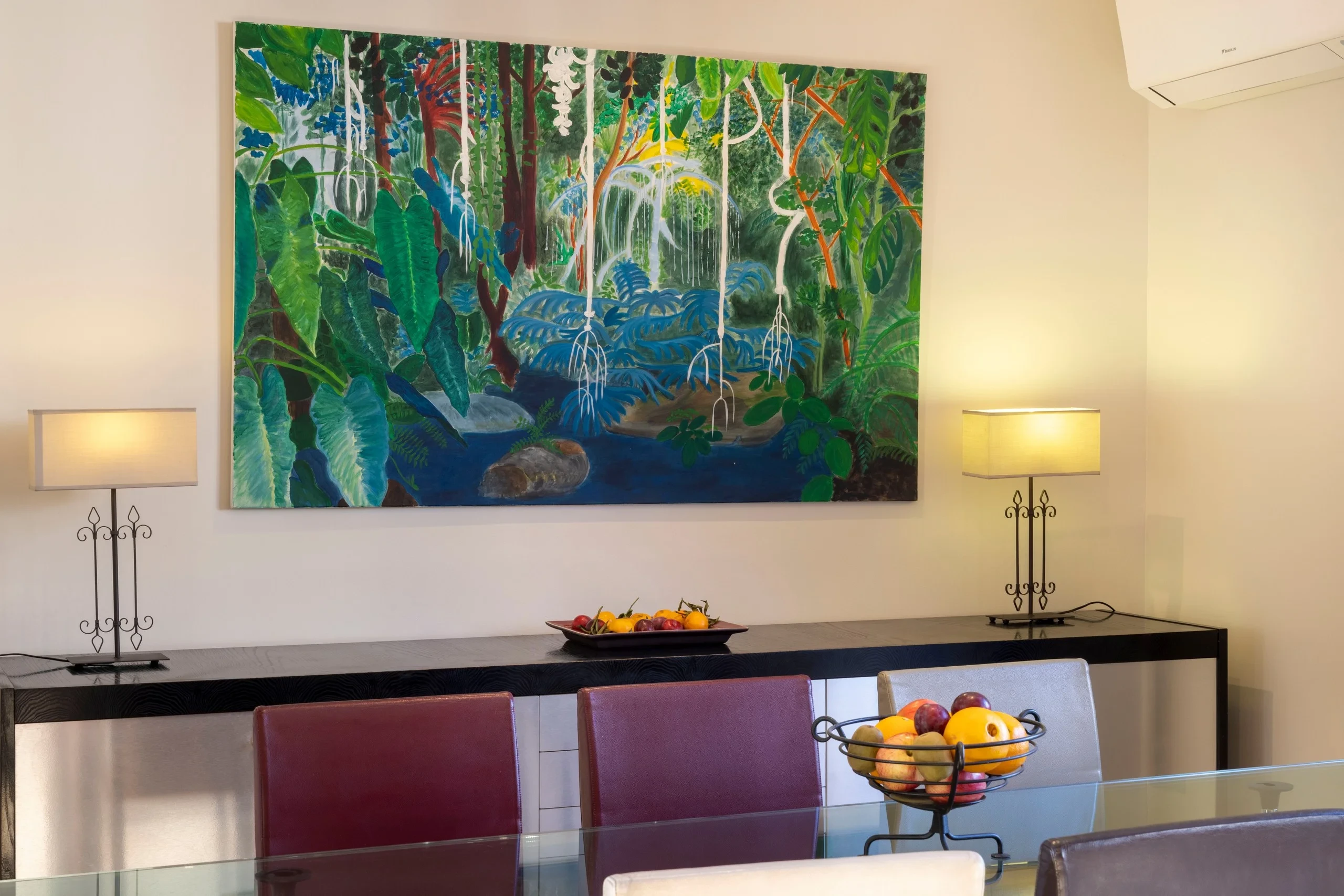 Salle à manger avec table en verre et grand tableau représentant une végétation tropicale au-dessus d’un buffet.