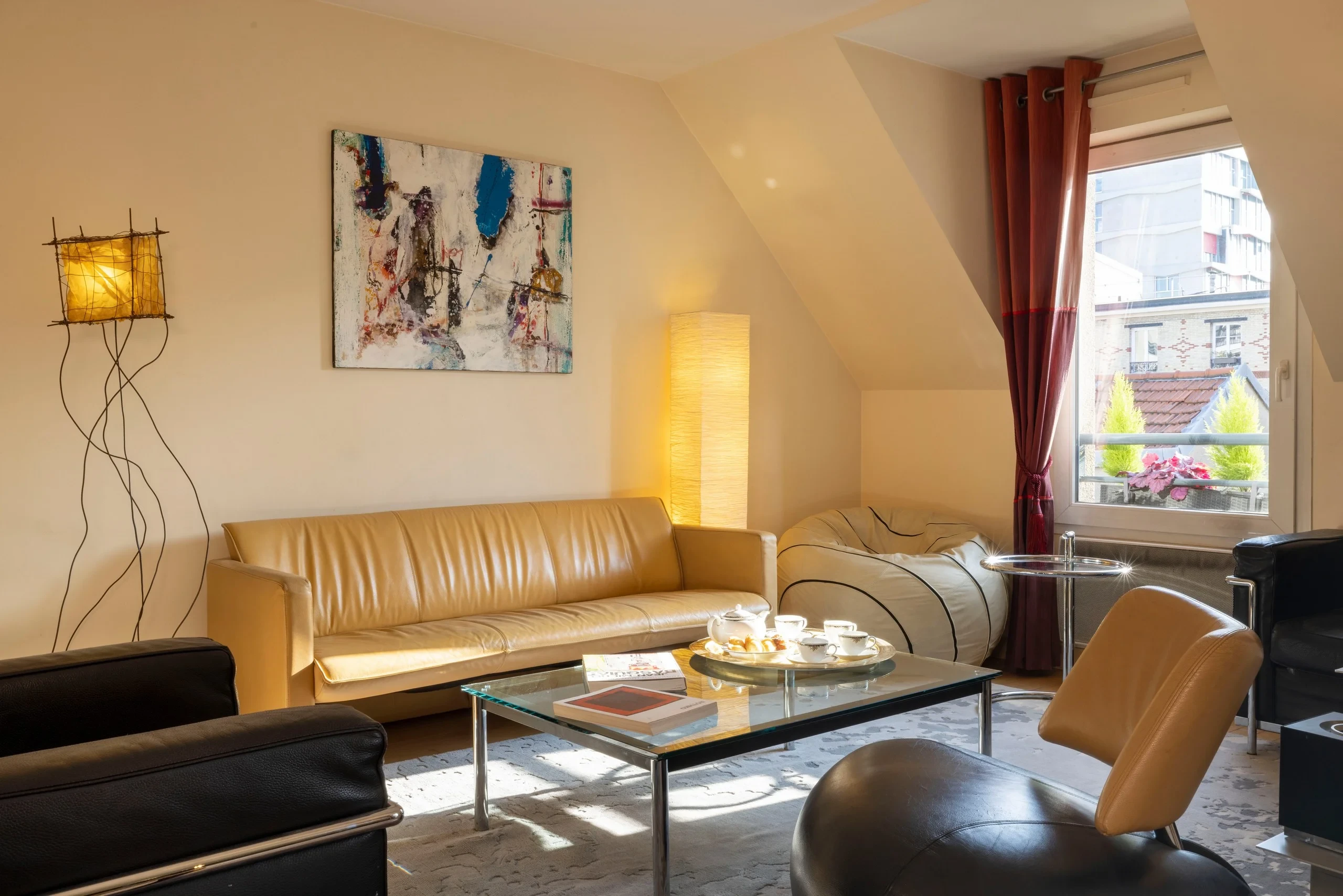 Salon avec canapé en cuir beige et fauteuils autour d’une table basse en verre, près d’une grande fenêtre.