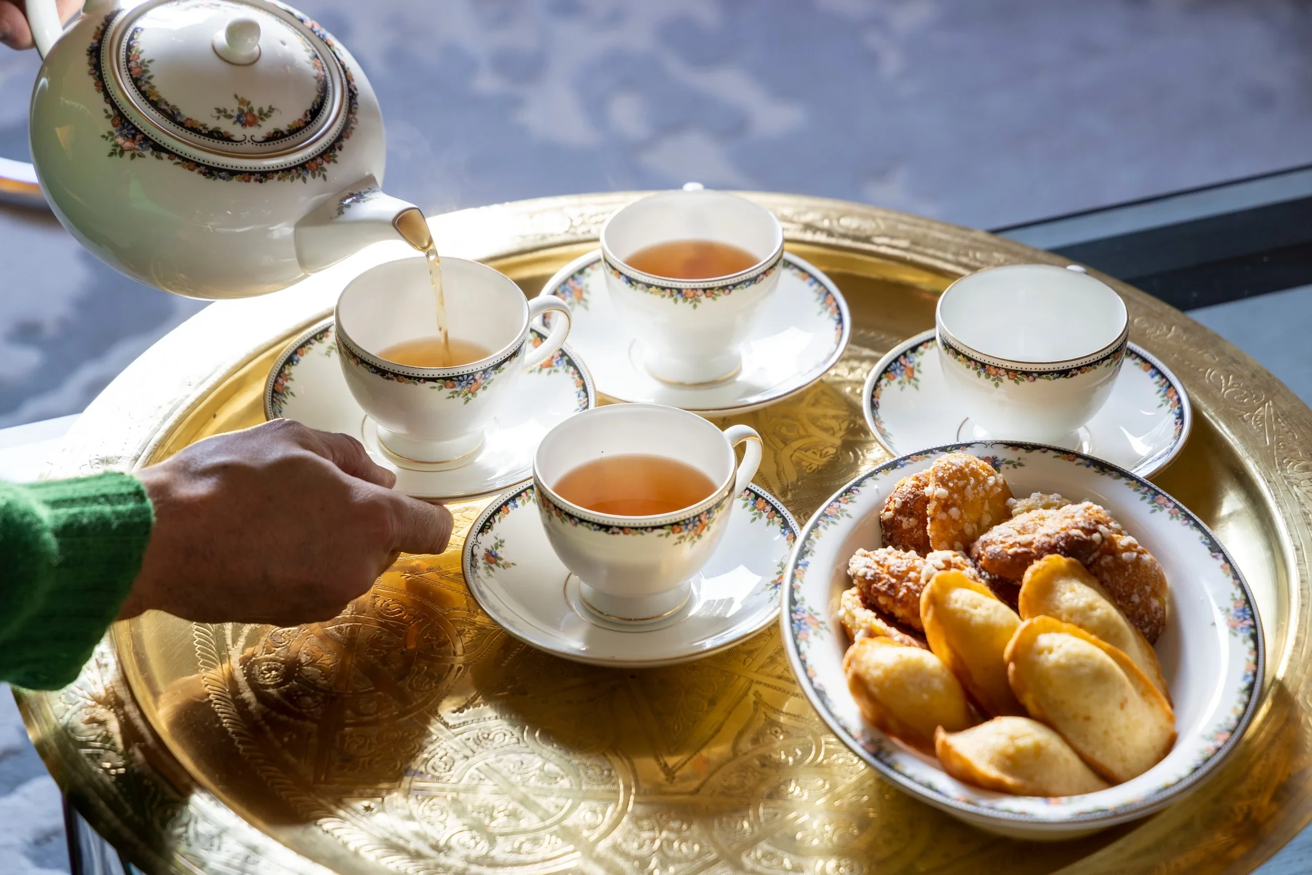Thé versé dans des tasses en porcelaine à motifs floraux, avec des pâtisseries sur un plateau doré.