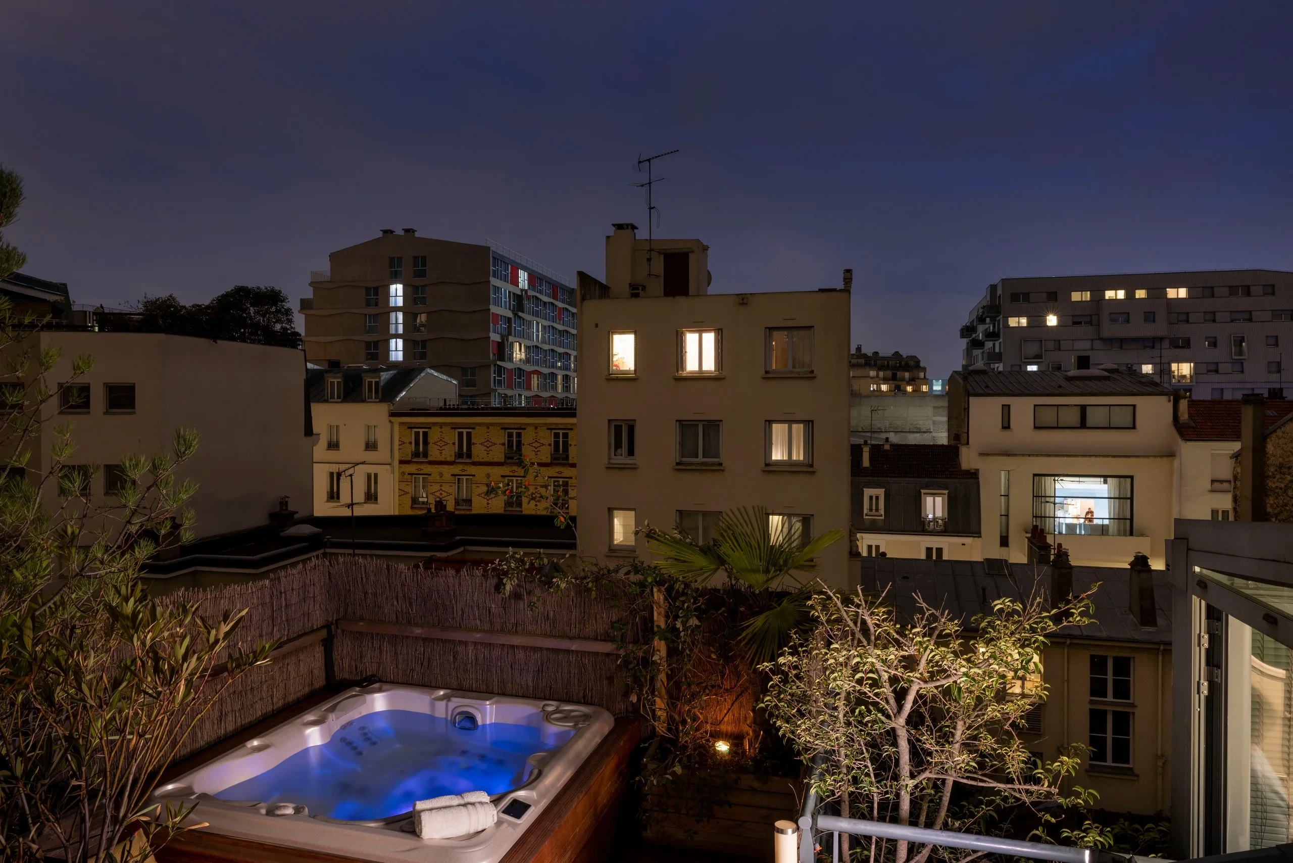 Bain à remous éclairé de bleu sur la terrasse végétalisée de MAISON LAVAUD - Paris, face aux immeubles voisins à la nuit tombée.