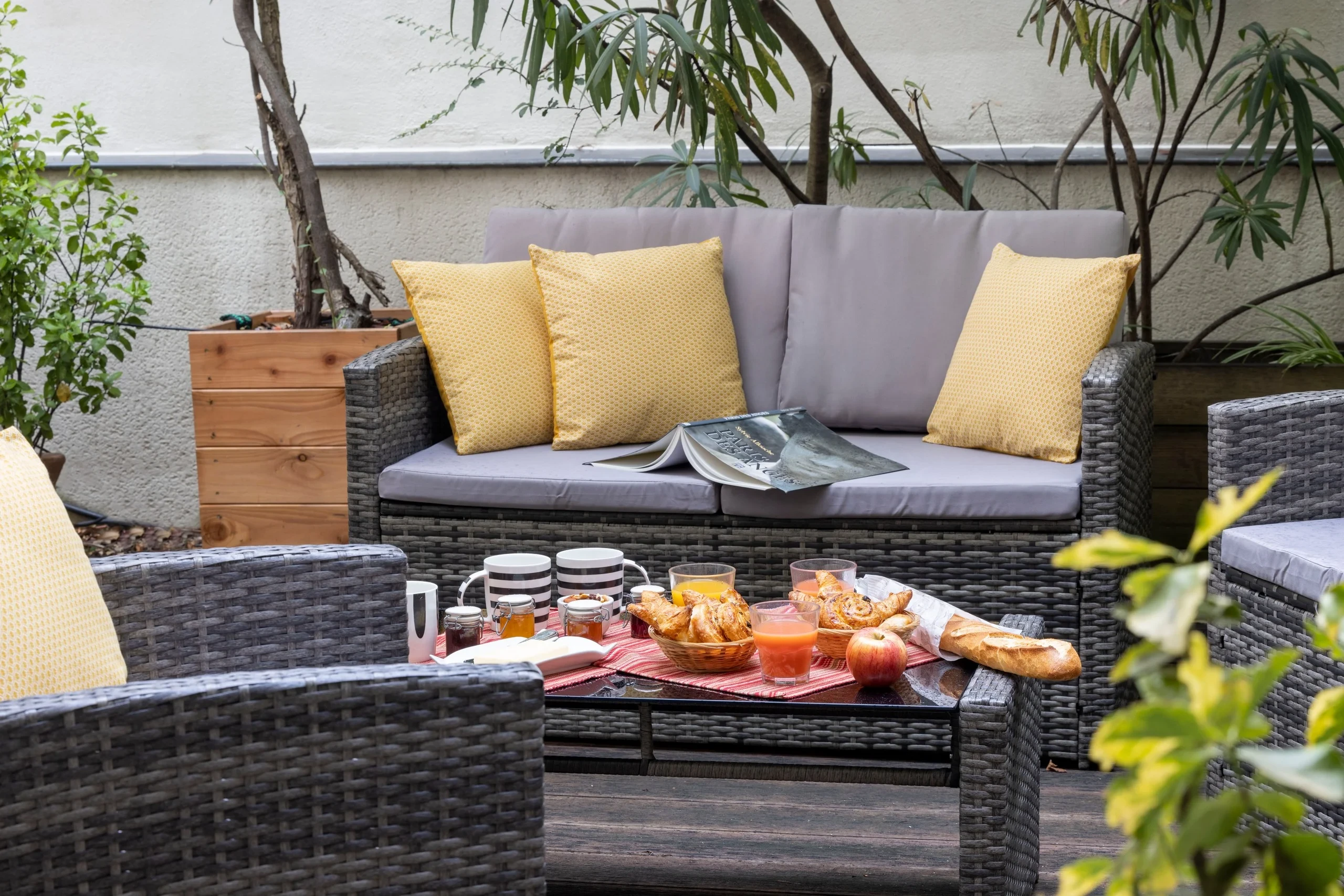 Petit-déjeuner sur une table basse devant un canapé de jardin gris aux coussins jaunes, entouré de végétation.