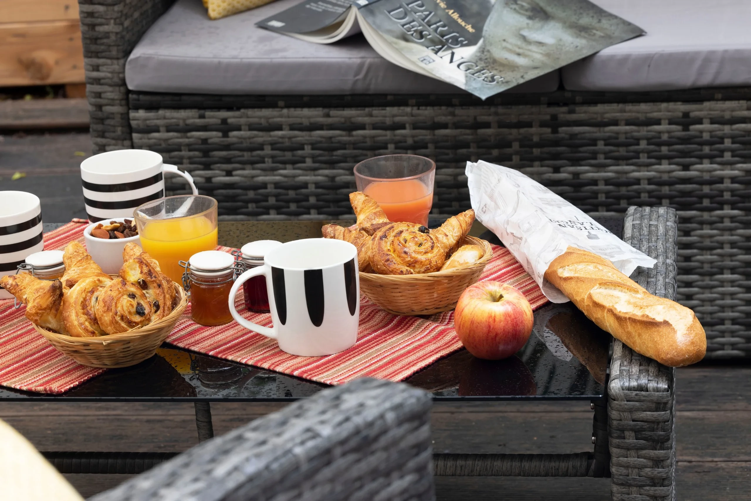 Petit-déjeuner composé de viennoiseries, de baguette, de confitures et de jus de fruits sur une table de jardin.