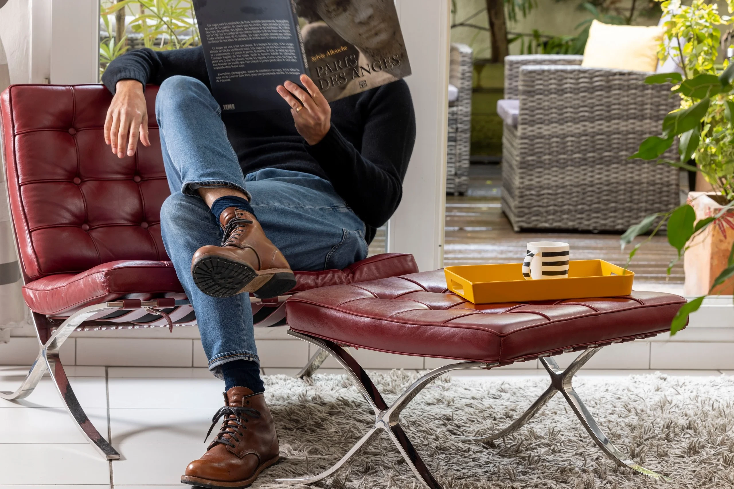 Une personne lit un livre dans un fauteuil en cuir bordeaux, près d’une baie vitrée donnant sur une terrasse végétalisée.