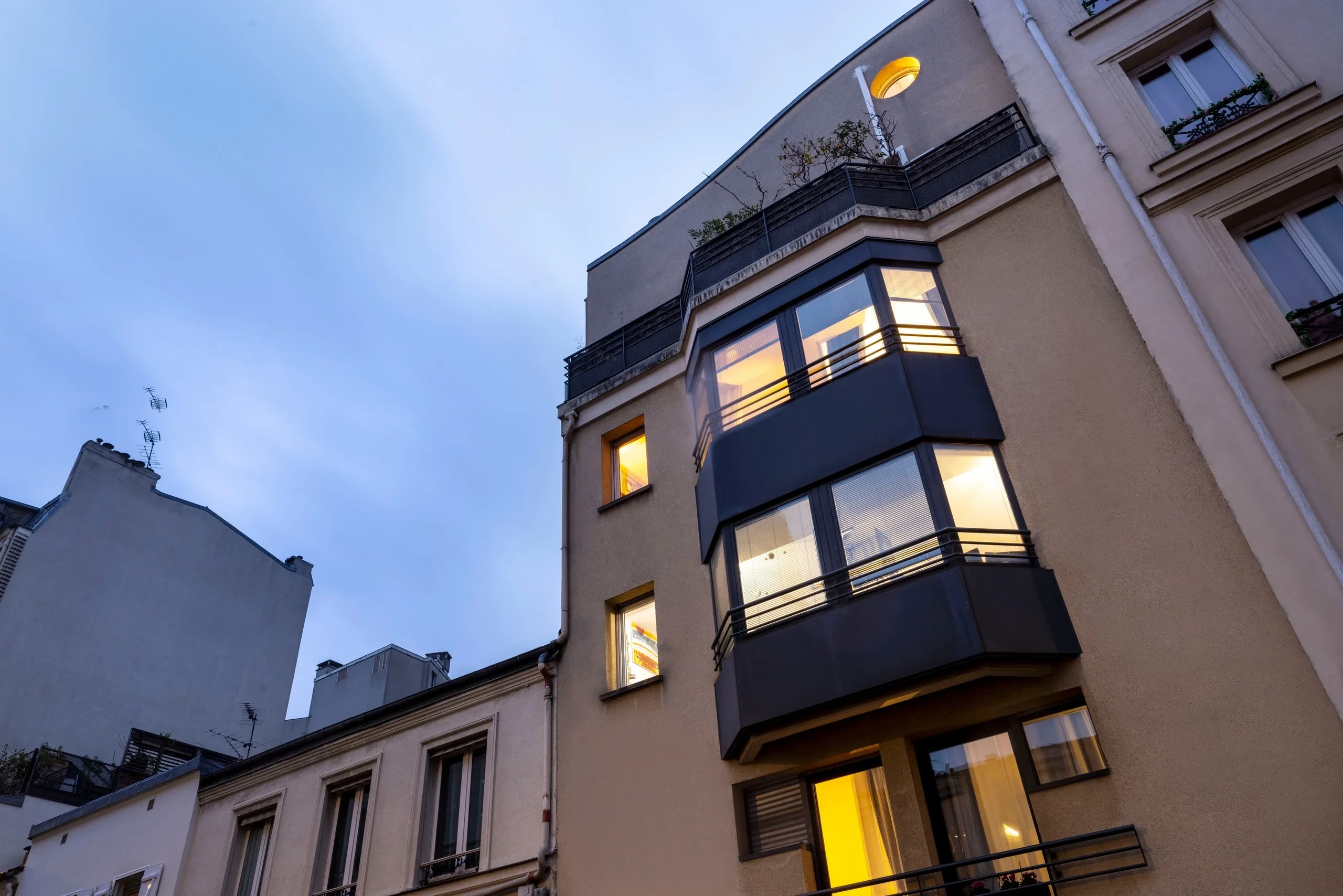 Façade de MAISON LAVAUD - Paris, avec des baies vitrées en saillie et des fenêtres éclairées à la tombée du jour.