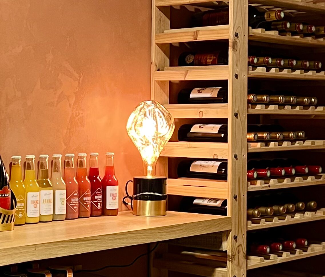Bouteilles de vin rangées dans des casiers en bois, près d’une lampe allumée et de bouteilles de jus de fruits.