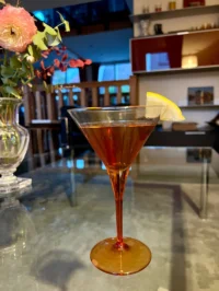 Cocktail ambré dans un verre à pied évasé, garni d’un quartier de citron et posé sur un comptoir en verre.