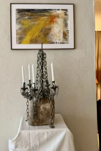 Vue de face d’un chandelier en chaînes métalliques sur une base massive, sous un tableau abstrait aux traits jaunes, noirs et orangés.