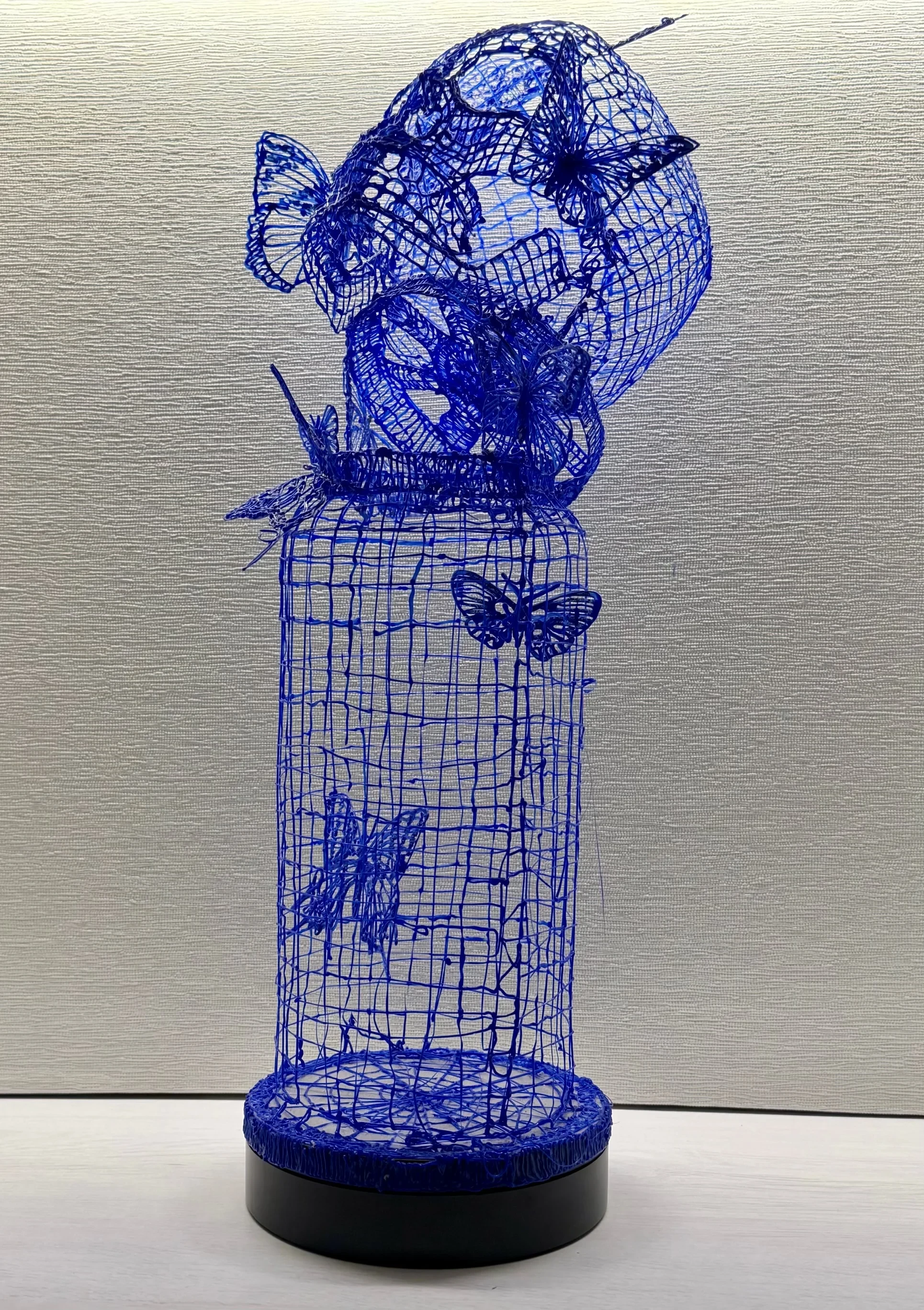 Sculpture bleue ajourée représentant un crâne entouré de papillons, sur un support cylindrique grillagé.