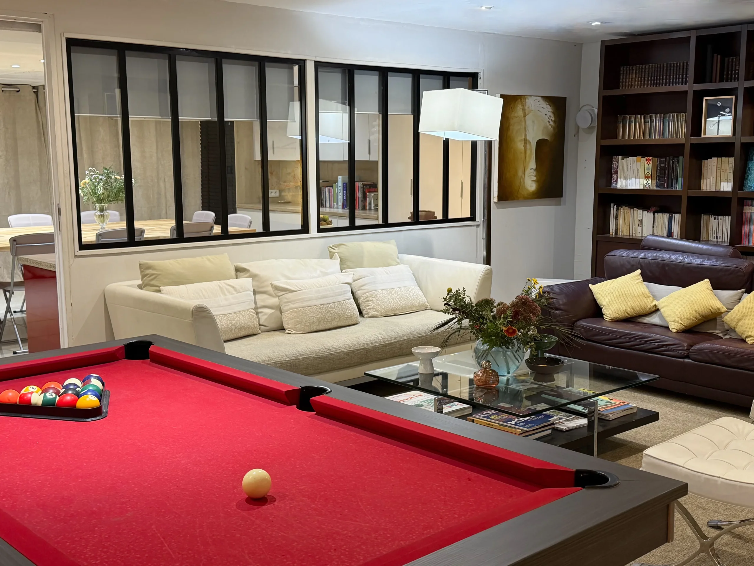 Billard à tapis rouge devant deux canapés, une bibliothèque et une verrière intérieure.