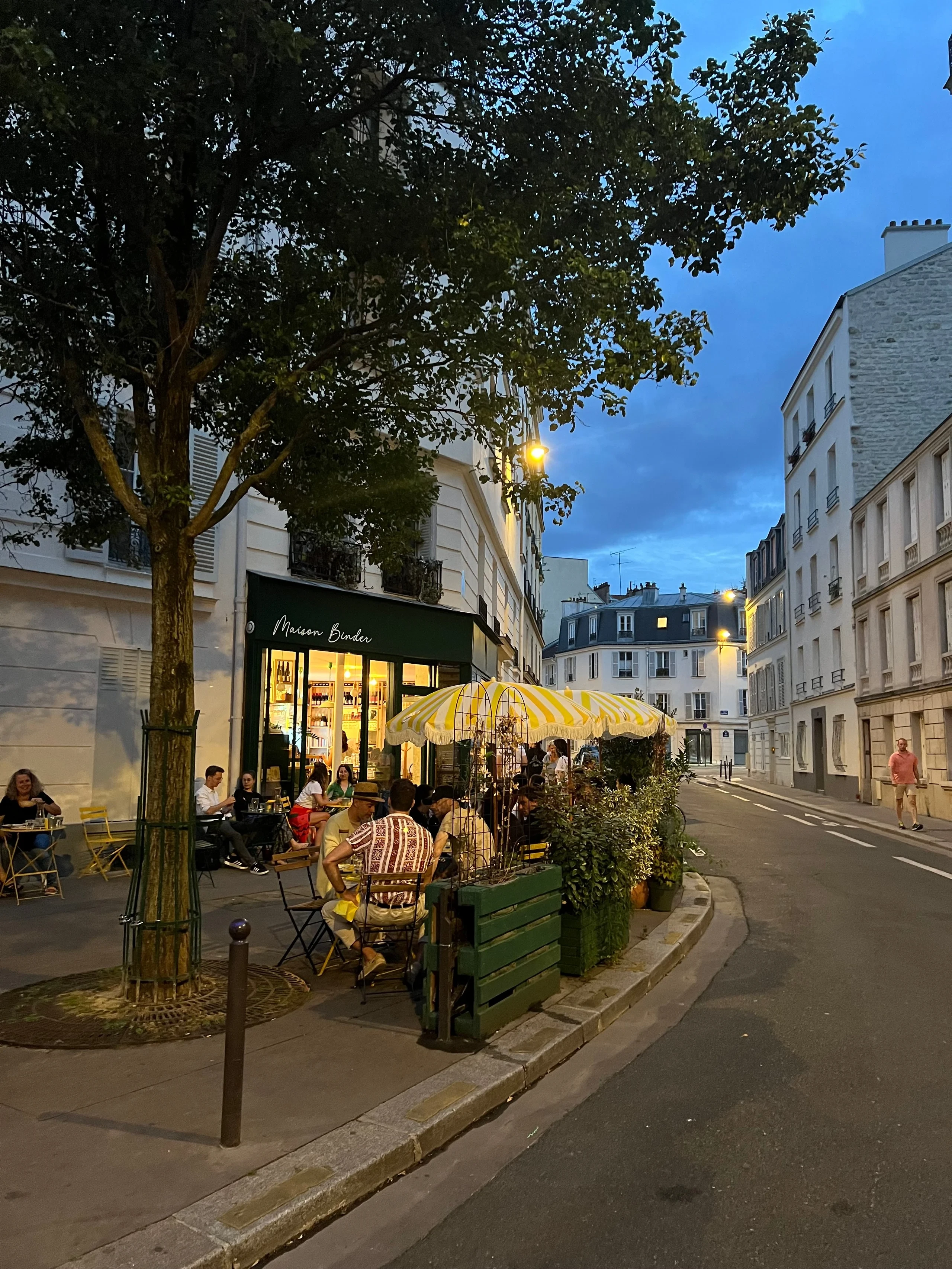Terrasse de la Maison Binder à un angle de rue au crépuscule, sous des parasols rayés jaunes et blancs.