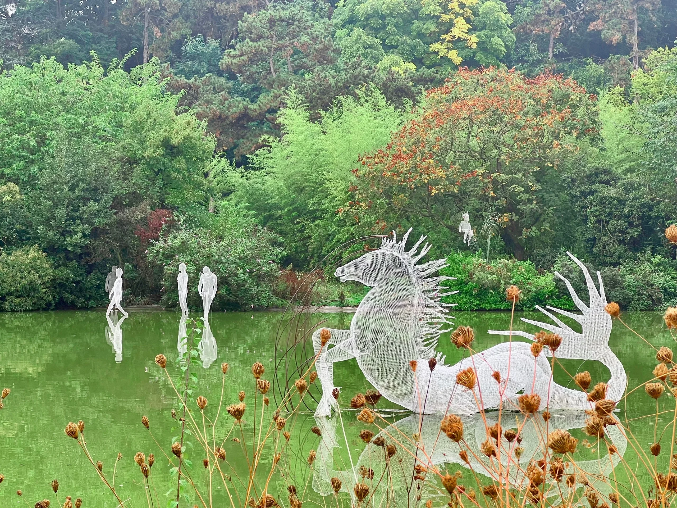 Sculpture blanche ajourée d’une créature fantastique dans un étang, avec des silhouettes humaines sculptées en arrière-plan.