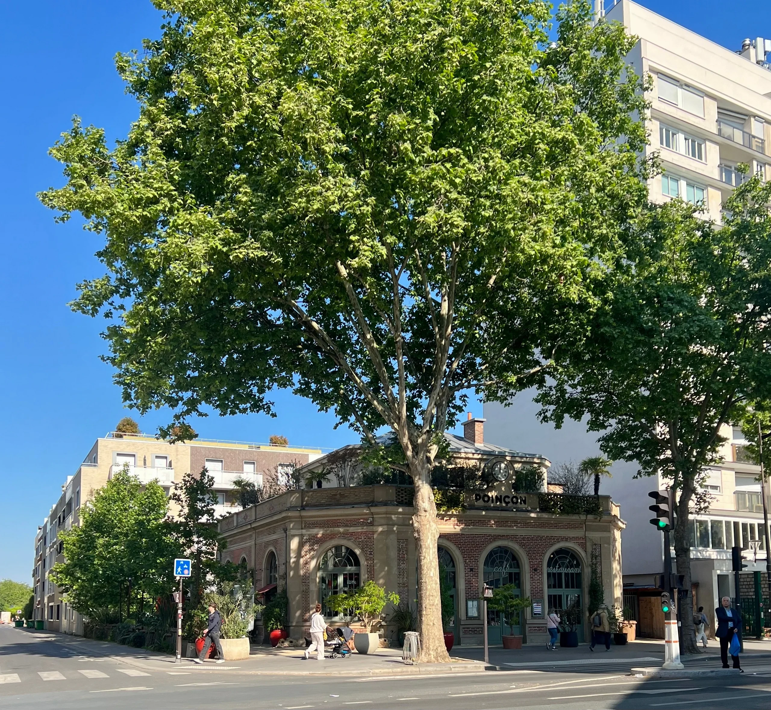 Façade en brique du café-restaurant Poinçon, avec de grandes fenêtres cintrées sous les arbres.