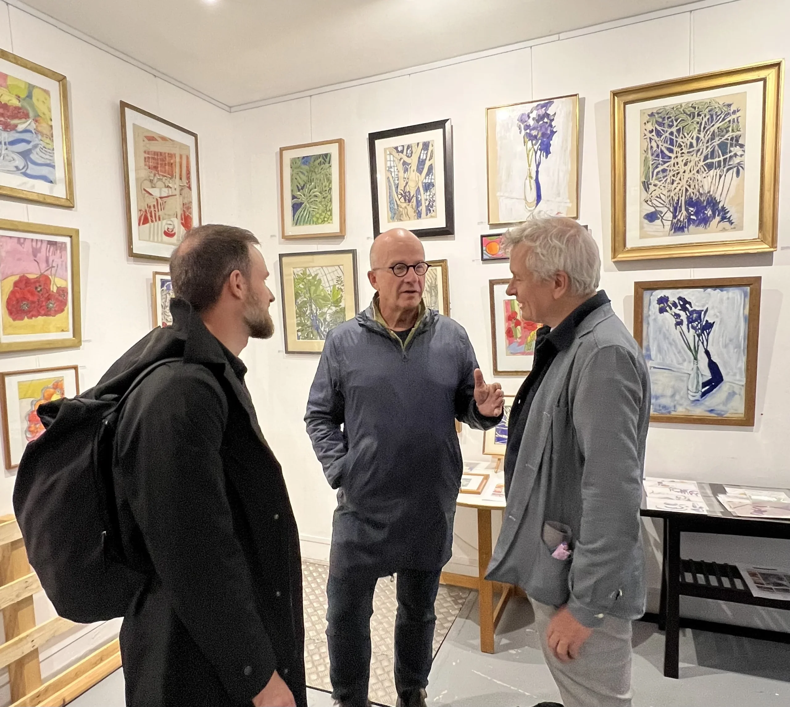Trois hommes discutent dans une salle d’exposition aux murs couverts de tableaux figuratifs et floraux.