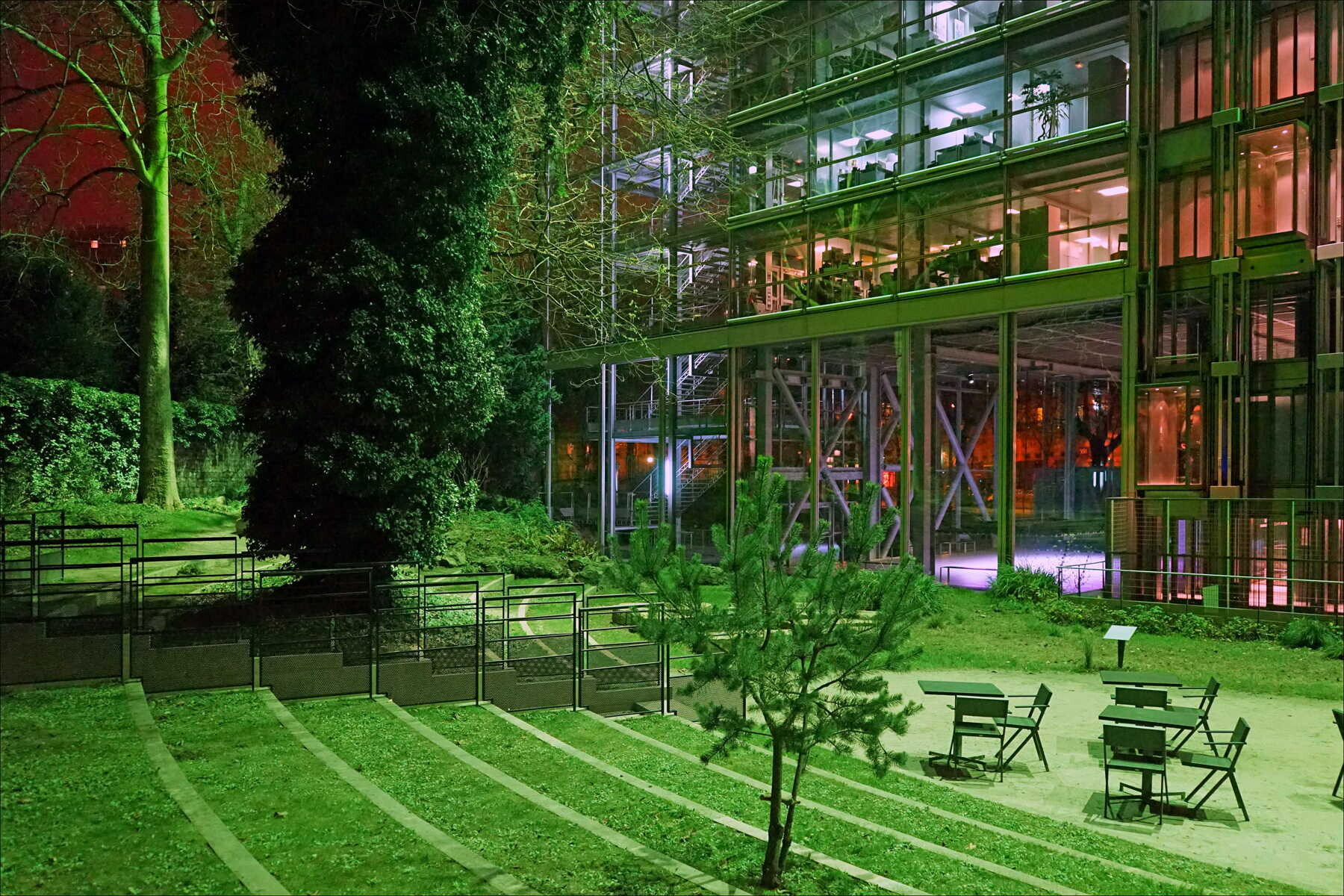 Jardin éclairé de nuit, avec des tables et des chaises devant une façade vitrée à plusieurs étages.